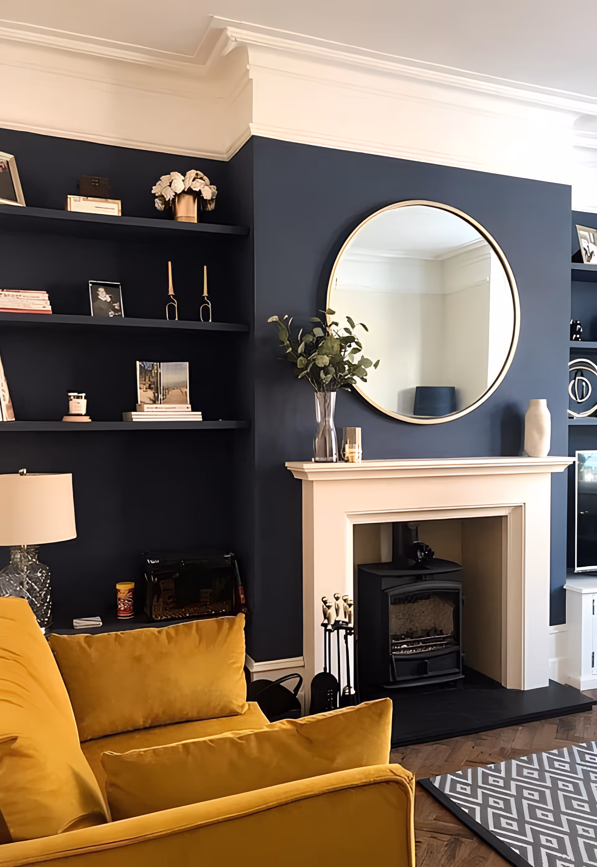 A cozy living room featuring a mustard yellow sofa, a white fireplace with a black stove insert, and a large round mirror above the fireplace. The walls are painted dark blue with built-in shelves holding decorative items such as books, candles, and vases. A patterned rug is partially visible on the wooden floor.