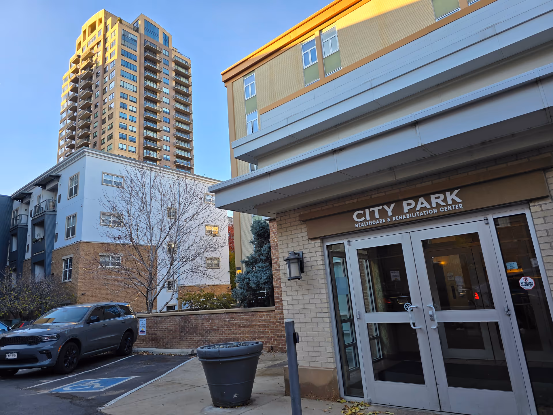 Exterior view of the entrance to City Park Healthcare & Rehabilitation Center with glass double doors. A large building and a tall apartment or office tower are visible in the background. There is a parking area with a gray SUV parked in a handicapped spot and a large empty planter near the entrance.