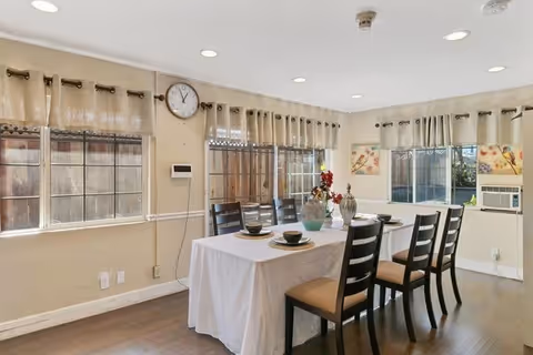 A bright dining room with a rectangular table covered with a white tablecloth and set with plates, bowls, and cups. Six dark wooden chairs with beige cushions surround the table. The room has large windows with beige curtains, a wall clock, and two framed floral paintings. The floor is wooden, and the ceiling has recessed lighting.