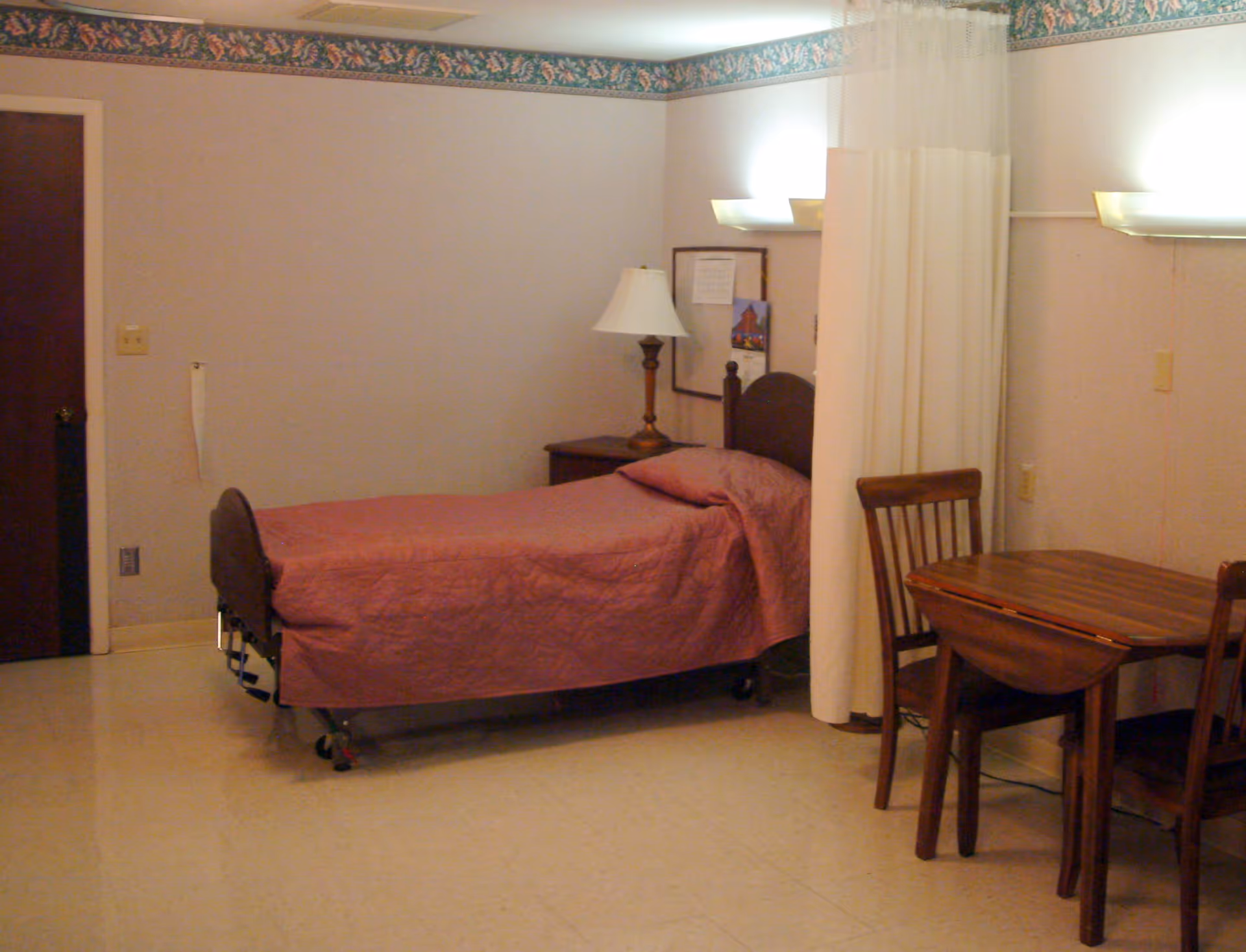 A simple room with a single hospital-style bed covered with a pink quilt, a wooden nightstand with a lamp, a small wooden table with two chairs, and a privacy curtain partially drawn around the bed. The walls are light-colored with a floral border near the ceiling.