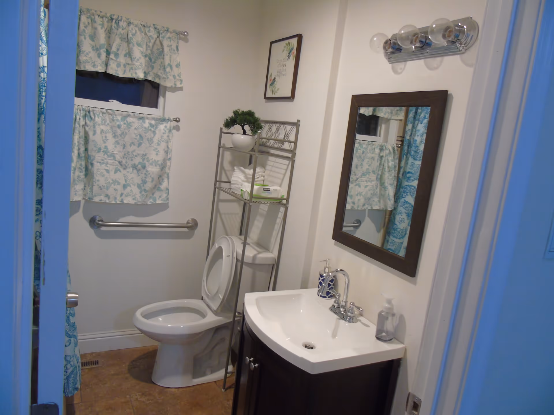 A small bathroom with a white toilet and a dark wood vanity with a white sink. Above the sink is a rectangular mirror with a dark frame and a light fixture with four bulbs. To the left of the toilet is a metal shelving unit holding folded white towels, a small potted plant, and a box of wipes. A window with floral curtains is above a grab bar on the wall. A blue and white patterned shower curtain is partially visible on the left side.