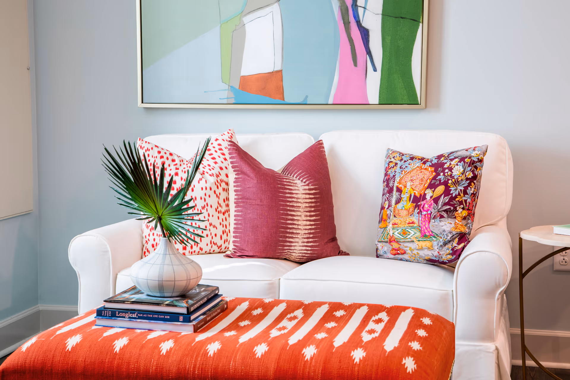 White loveseat with colorful throw pillows, an orange patterned ottoman topped with books and a vase, and an abstract painting on the wall behind.