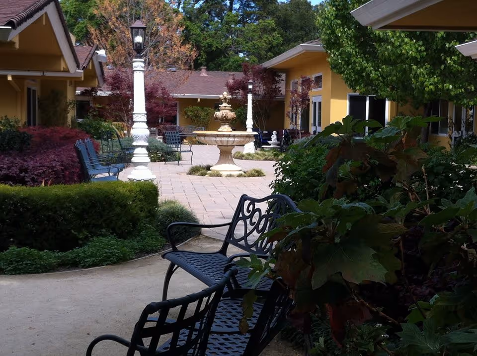 Outdoor courtyard area with black metal benches, a stone fountain in the center, surrounded by yellow buildings, green bushes, and trees with some autumn-colored leaves.