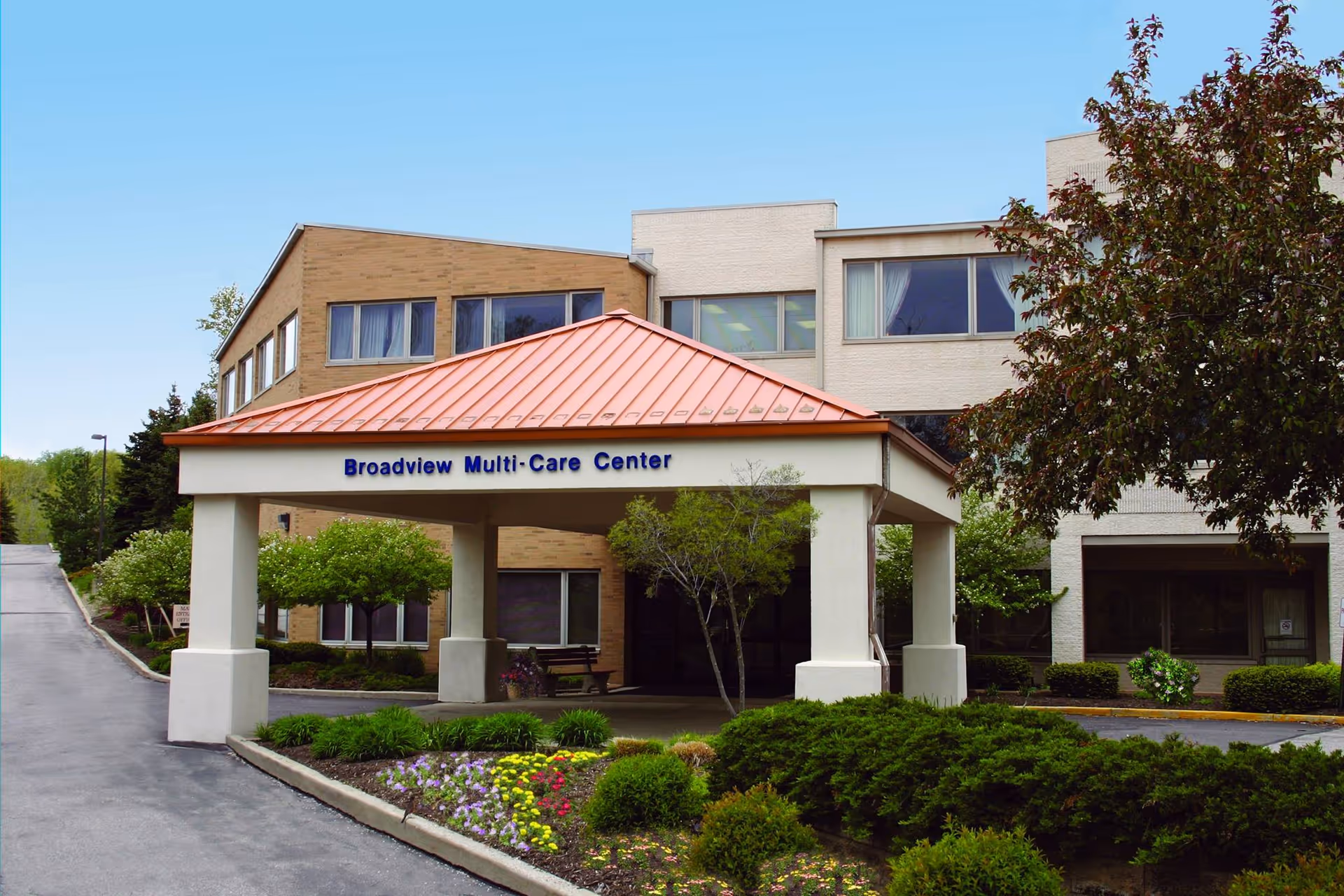 Exterior view of Broadview Multi-Care Center showing the entrance with a covered driveway, surrounded by landscaped bushes, flowers, and trees under a clear blue sky.