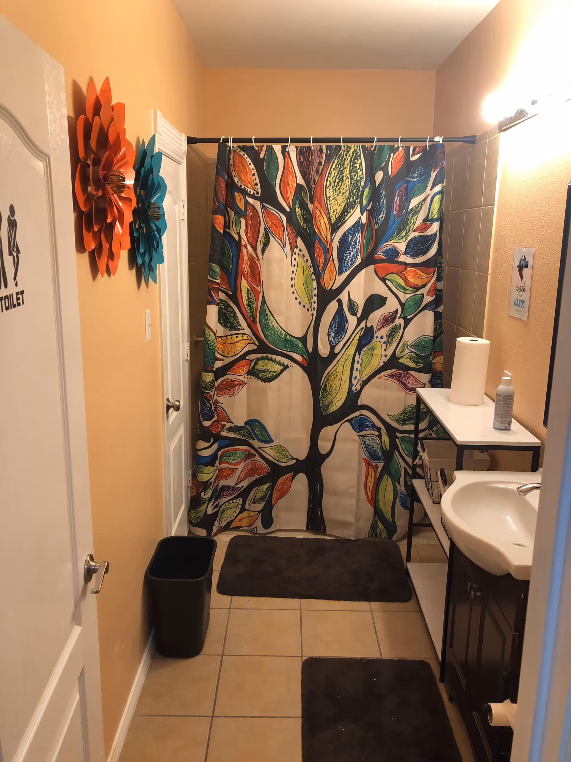 A bathroom with beige walls and tiled floor featuring a colorful tree-patterned shower curtain. There are two decorative flower wall hangings in orange and teal on the left wall. A white door with a toilet sign is on the left side. The bathroom has a dark brown vanity with a white sink, a small shelving unit holding paper towels and soap, and two dark brown bath mats on the floor. A black trash bin is placed near the door.