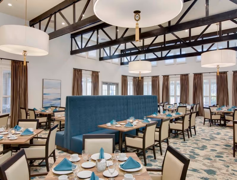 Spacious dining room with set tables and teal napkins, a blue tufted banquette, high exposed-beam ceiling, and large pendant lights.