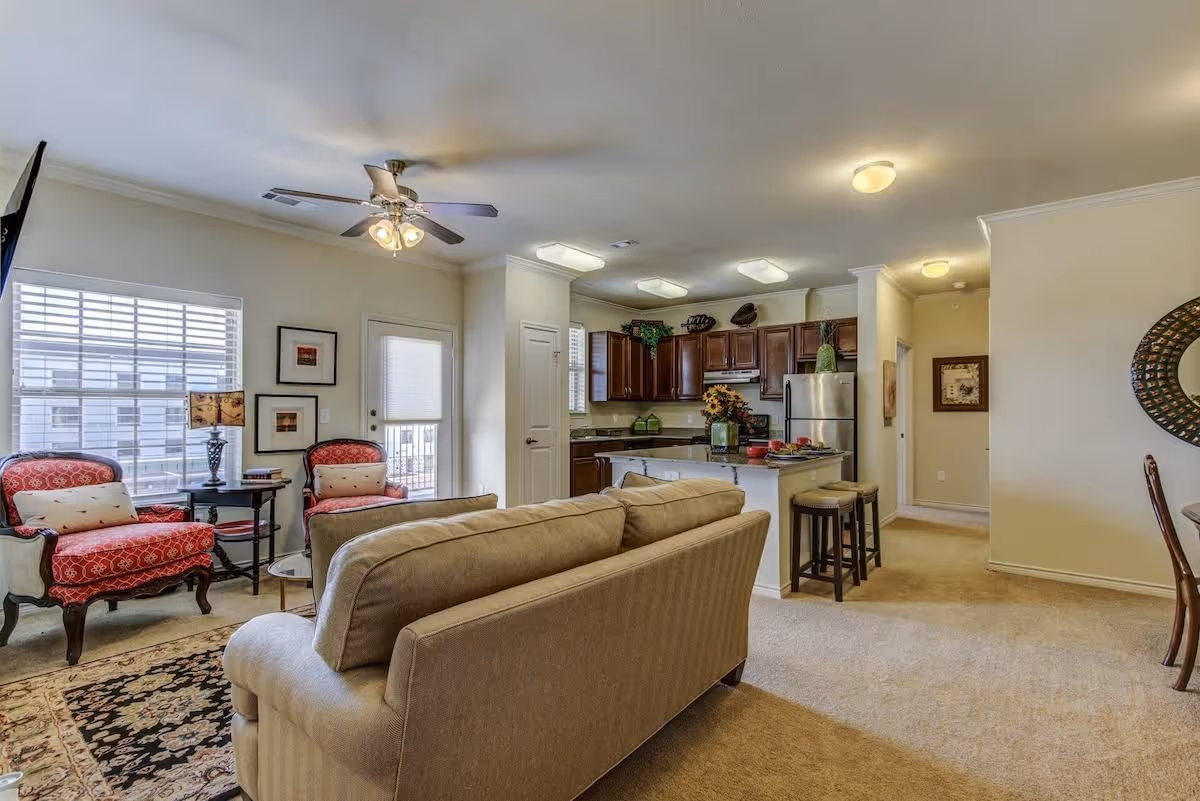 Open-plan living room with a beige sofa facing two red patterned armchairs by a window and a kitchen with a center island and dark wood cabinets in the background.