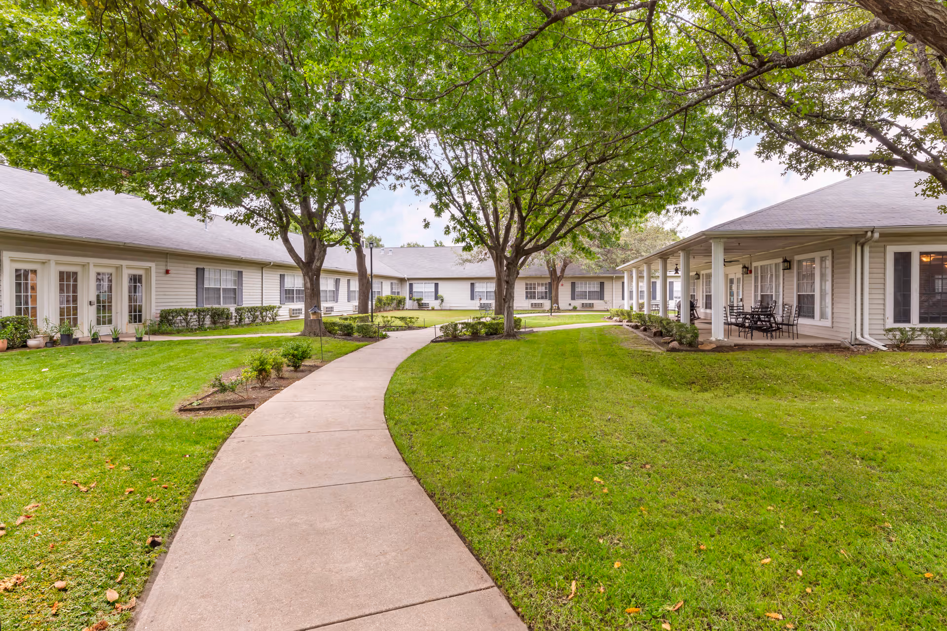 A curved concrete pathway runs through a well-maintained grassy courtyard with several large trees providing shade. Surrounding the courtyard are single-story buildings with light-colored siding, windows, and some outdoor seating areas under covered porches.