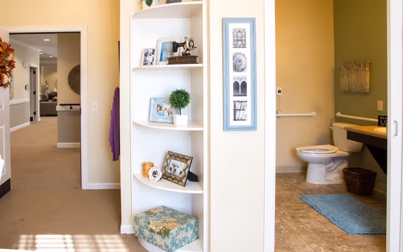 Interior view showing a hallway leading to a living area, with a white corner shelf displaying framed photos, a small plant, and decorative items. To the right, there is a bathroom with a toilet, a blue bath mat, a wicker basket, and a countertop with a sink. The walls are painted light yellow and beige.