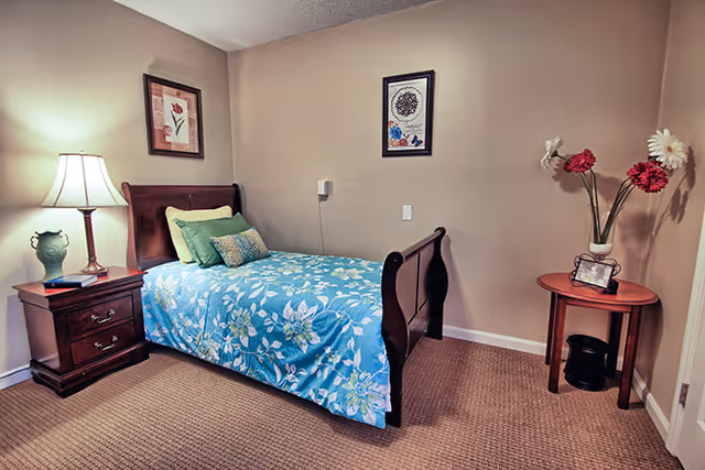 A small bedroom with a single wooden bed covered with a blue floral bedspread and green pillows. There is a wooden nightstand with a lamp and books on the left side of the bed. On the right side, there is a round wooden table with a vase holding three large flowers and a small framed picture. The walls are beige with two framed pictures hanging above the bed.