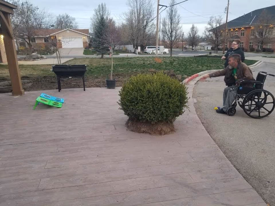 An outdoor patio area with a wooden deck, a green bush planted in the deck, a black raised planter, and a colorful cornhole game set. In the background, a man in a wheelchair is throwing a bean bag while another person stands behind him. There are houses, trees, and a parking lot visible further back.