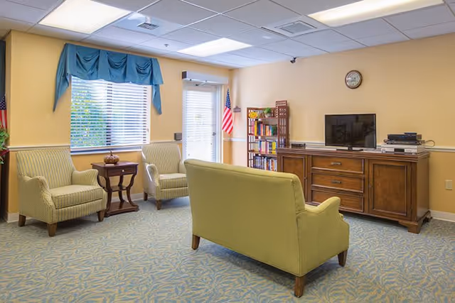 A cozy living room with two armchairs and a loveseat upholstered in light green fabric, a wooden side table with a decorative item, a wooden media console with a flat-screen TV and electronic devices, a bookshelf filled with books, an American flag near a door with window blinds, and a window with blue valance curtains letting in natural light.
