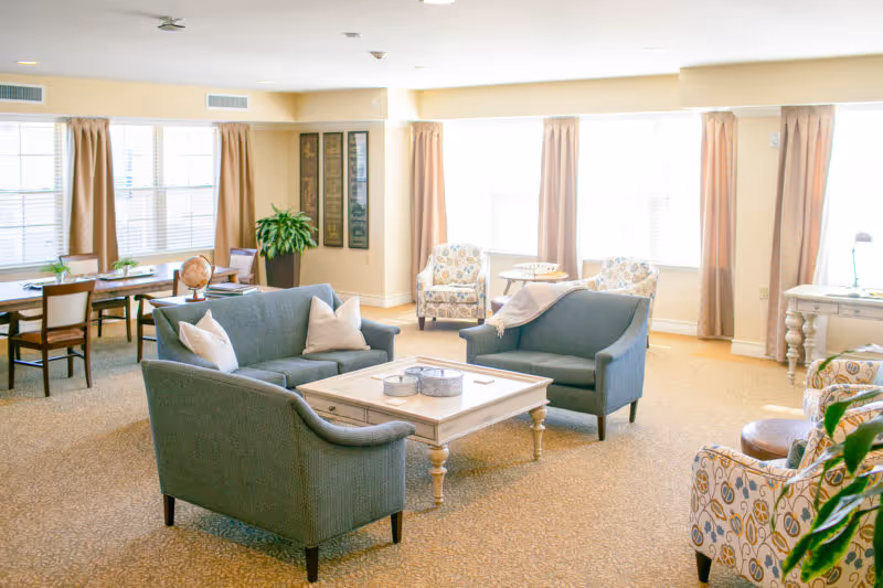 Bright senior living common room with blue sofas, patterned armchairs, a central coffee table and a dining table by large windows.