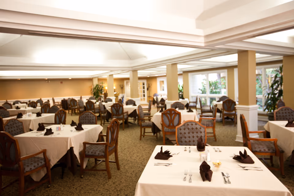 Spacious dining room with multiple tables set with white tablecloths, chairs, and folded napkins.