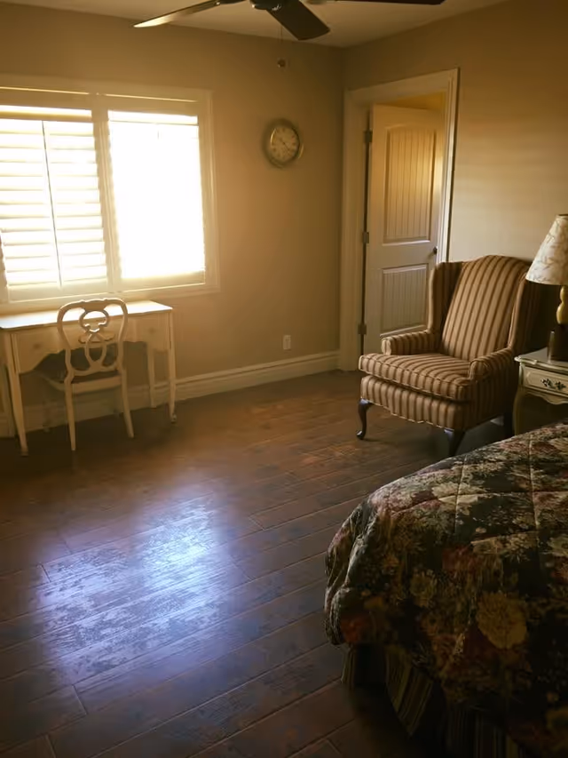 A bedroom with wooden flooring featuring a floral-patterned bedspread on the bed, a striped upholstered armchair, a small white desk with a matching chair near a window with white shutters, a wall clock, and a ceiling fan.