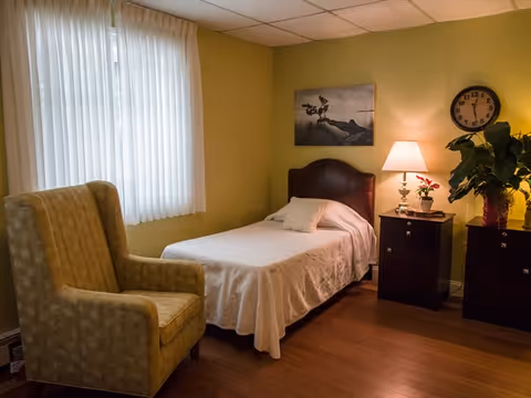 A cozy bedroom with a single bed covered in a white bedspread, a cushioned armchair, two dark wooden nightstands with a lamp, a clock on the wall, a potted plant, and a framed picture above the bed. The room has light green walls and a window with white curtains.