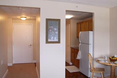 Interior view of a senior living apartment showing a hallway entry, a small kitchenette with refrigerator and cabinets, and a round dining table with chairs.