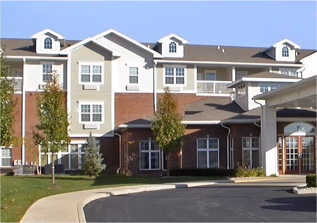 Exterior view of a multi-story senior living facility building with brick and light-colored siding, several windows, small trees, and a curved driveway in front.