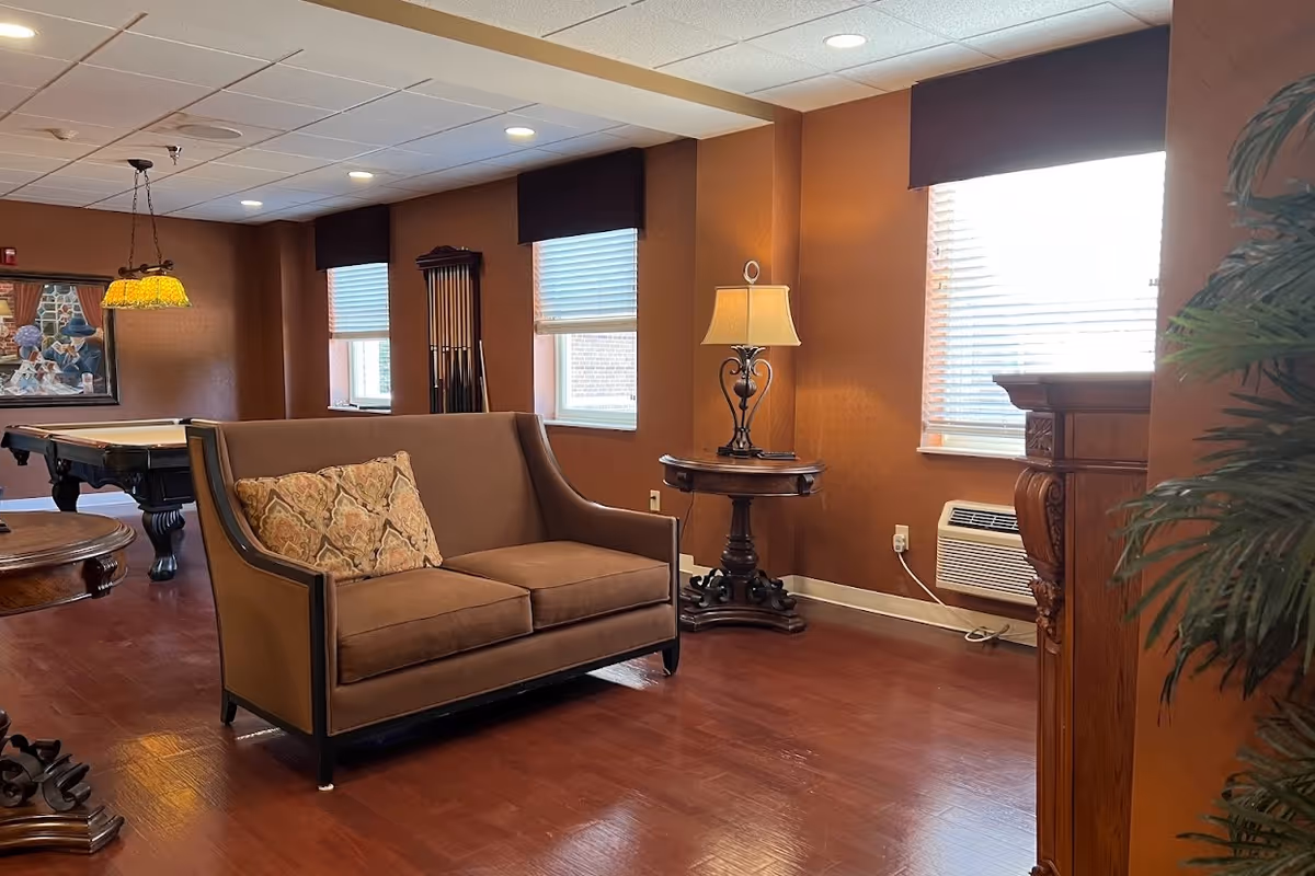 A warm-toned lounge with a brown loveseat, side table and lamp, a pool table in the background, and windows with blinds.