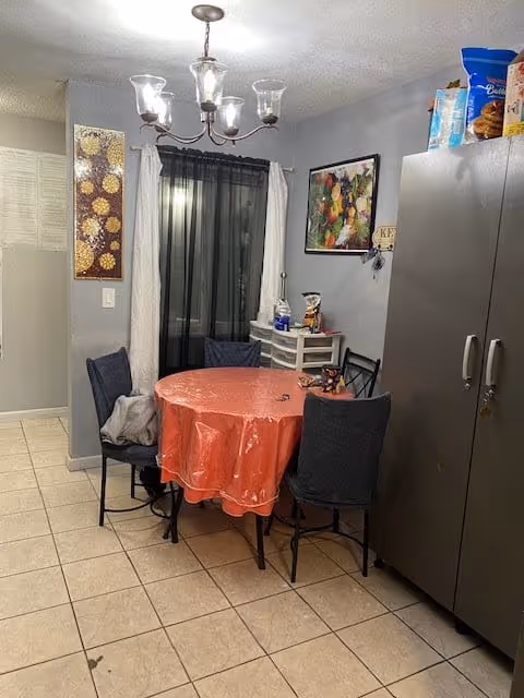 A small dining area with a round table covered by a red plastic tablecloth surrounded by four dark-colored chairs. There is a chandelier with five lights hanging above the table. Behind the table is a window with white curtains and a black sheer curtain. On the right side, there is a tall gray cabinet with various food items on top. The walls are painted gray and decorated with two framed pictures and a small shelf holding some items.