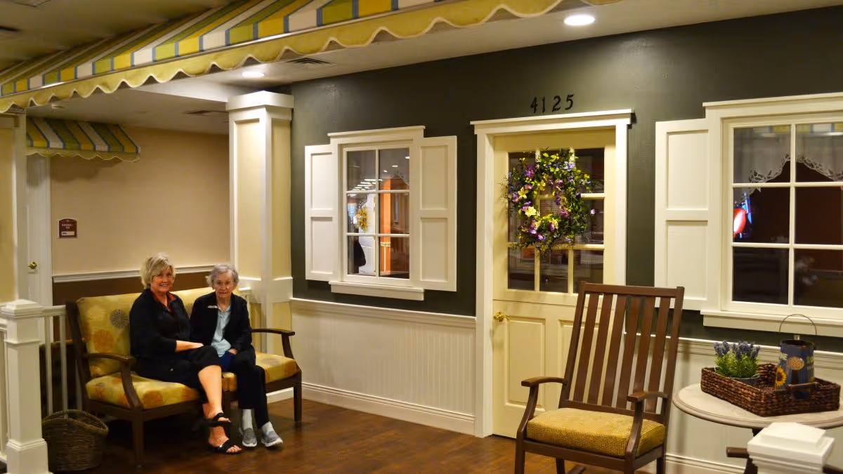 Two women sitting on a cushioned bench in a cozy indoor setting designed to look like a home exterior, with a door decorated with a floral wreath, windows with shutters, a wooden rocking chair, and a small table with a basket and decorative items.