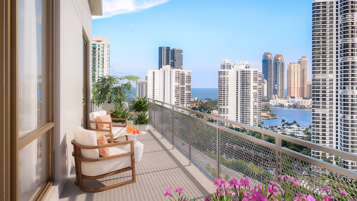 A sunlit balcony with cushioned chairs, potted plants and a railing overlooking high-rise buildings and water.