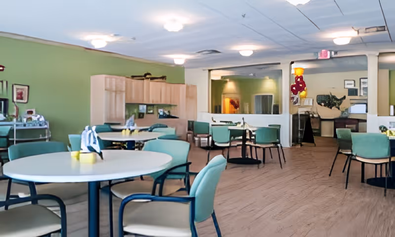 A bright communal dining area with round tables and teal chairs in a senior living facility.