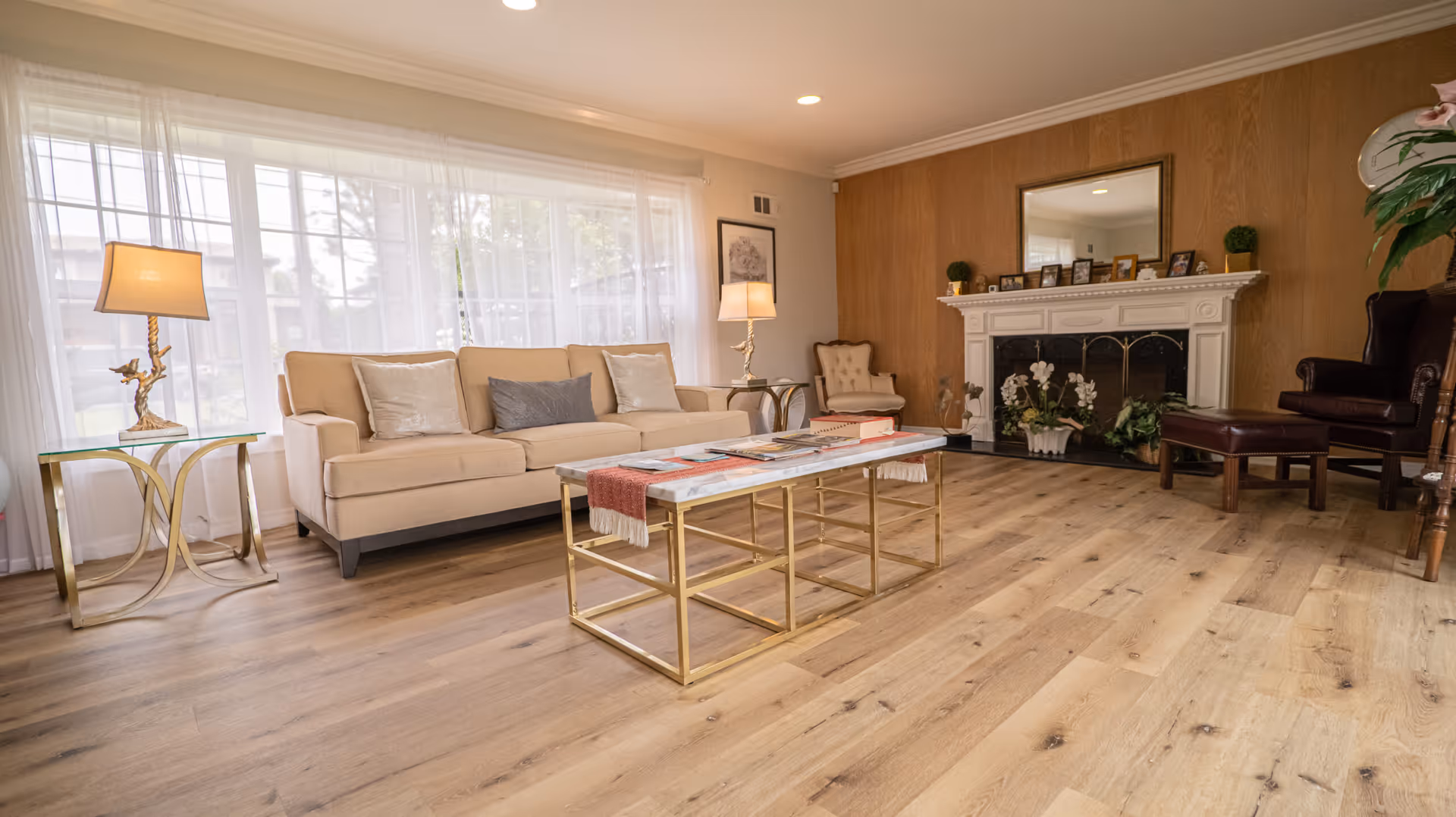 Bright living room with a beige sofa, gold-accent coffee table, fireplace, and wide wood-plank flooring.