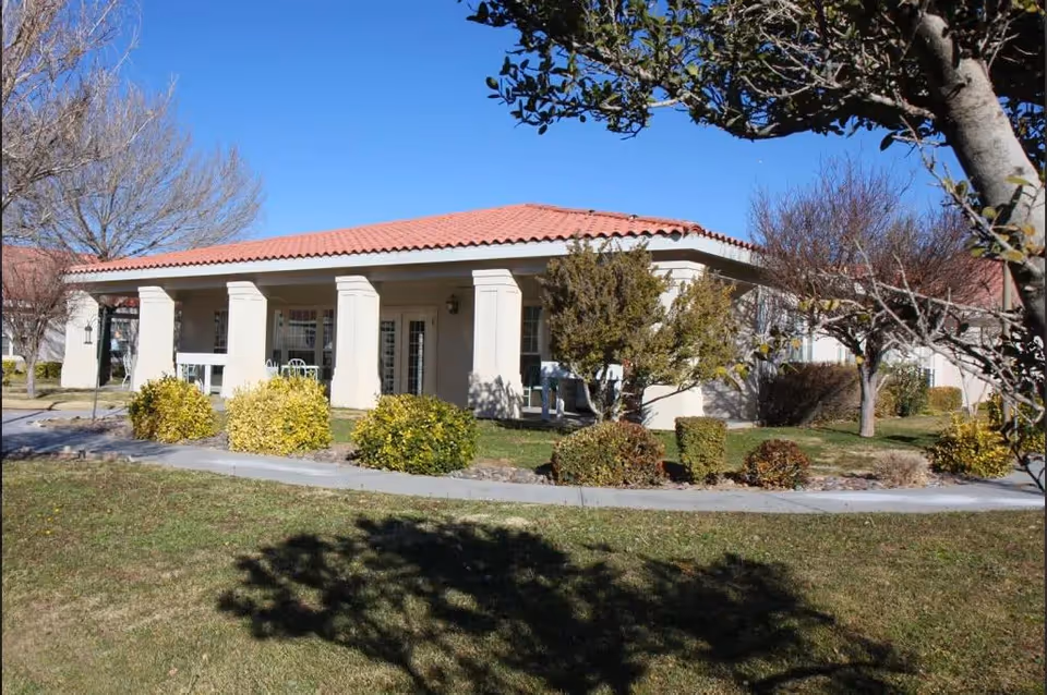 Single-story building with a red tiled roof and white columns on the porch, surrounded by bushes and trees with a clear blue sky in the background.
