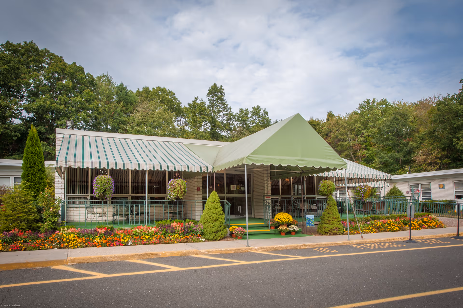 Front entrance of a single-story senior living facility with green striped awnings, hanging flower baskets, and colorful flower beds.