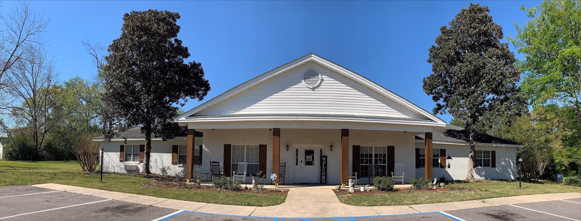 Front exterior view of a single-story assisted living facility building with white brick walls, brown shutters, and a covered porch with rocking chairs. Two large trees flank the entrance, and there is a 'WELCOME' sign near the door. The sky is clear and blue, and there is a parking area in front with marked spaces.
