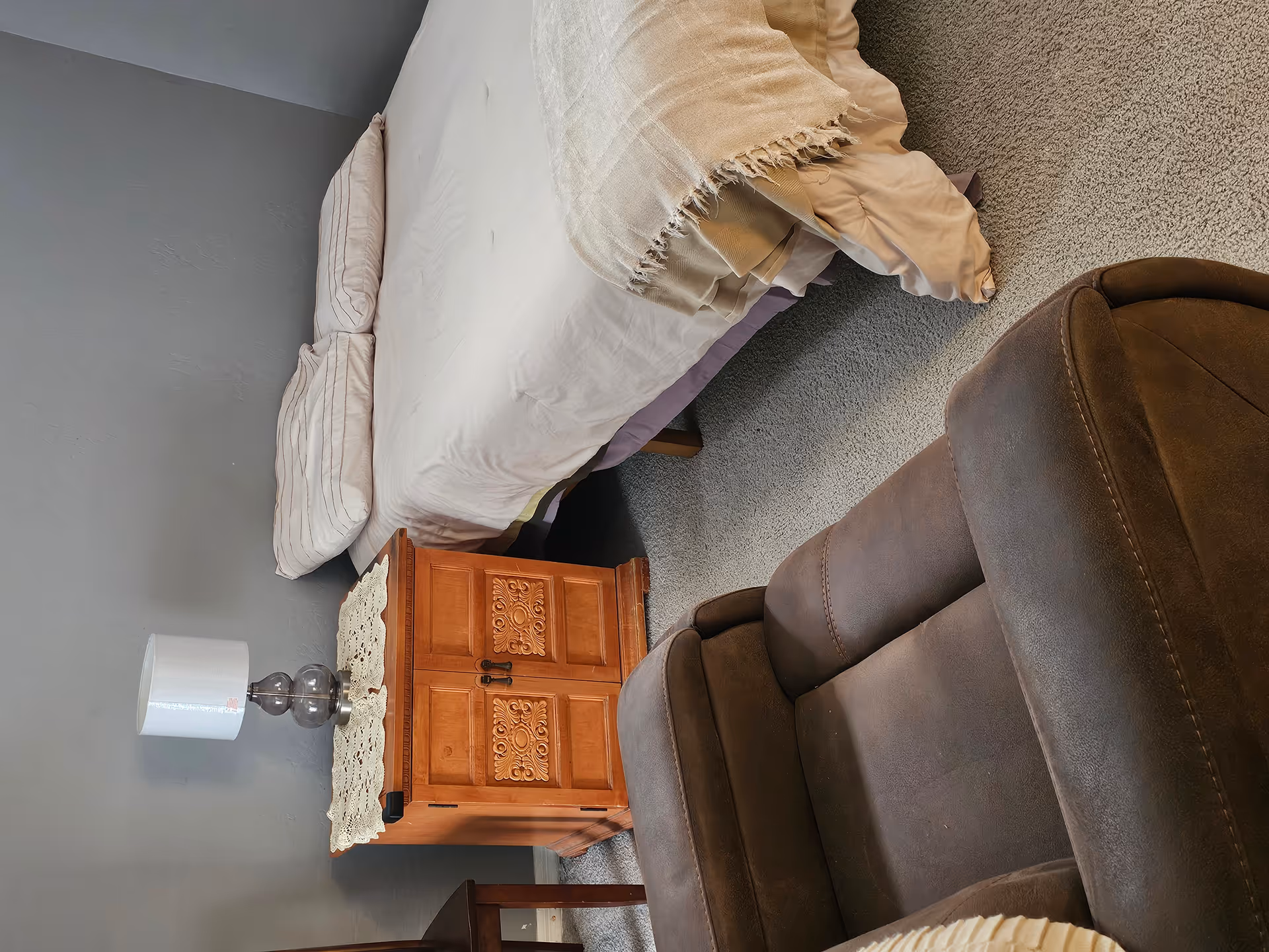 A bedroom corner with a bed, a carved wooden nightstand holding a lamp, and a brown recliner on carpet.