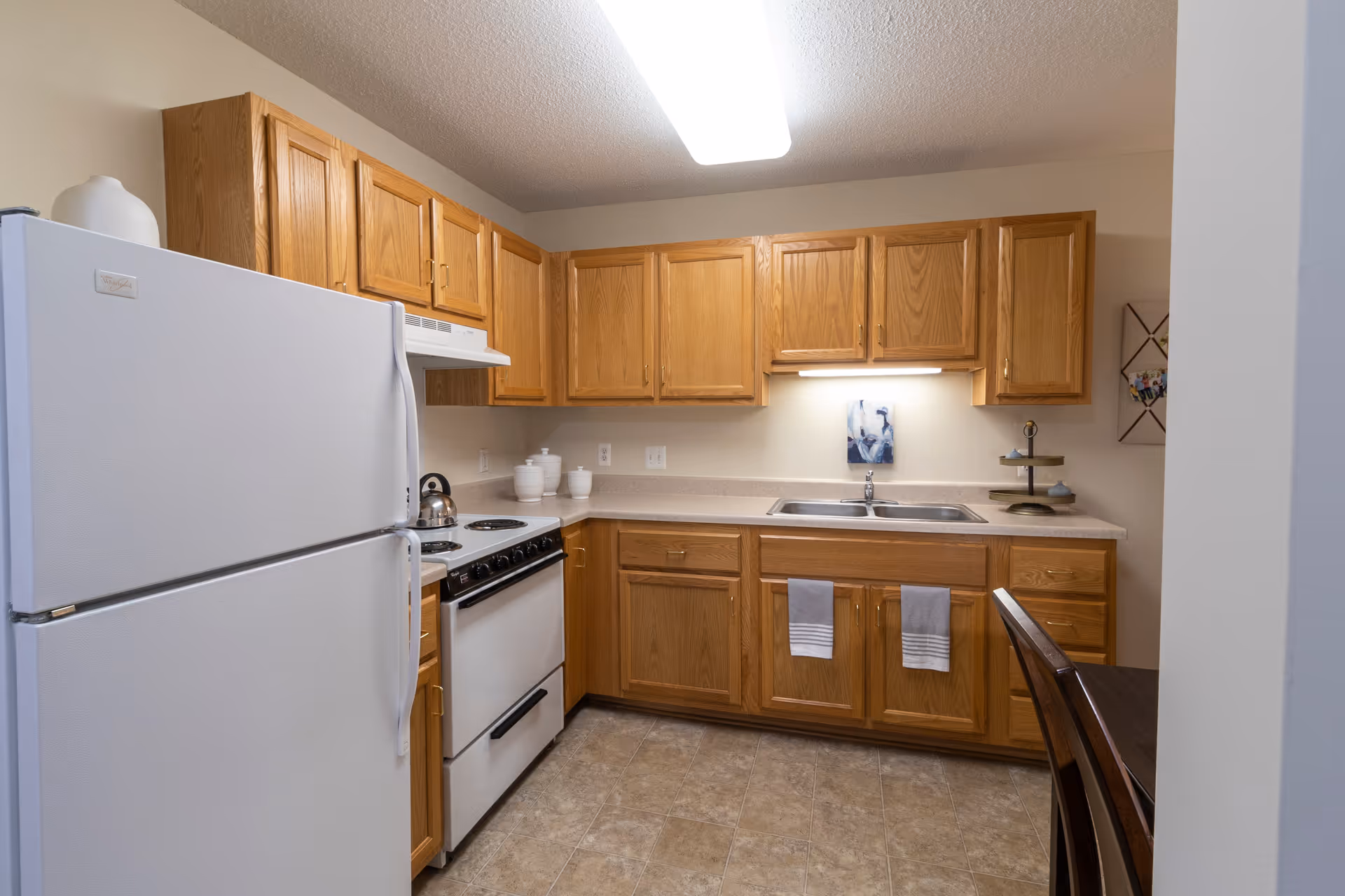 A kitchen with wooden cabinets, a white refrigerator, a white stove with a kettle on it, a double sink, and a countertop with decorative items. There are two towels hanging on the cabinet doors under the sink and a dining table with chairs partially visible on the right.