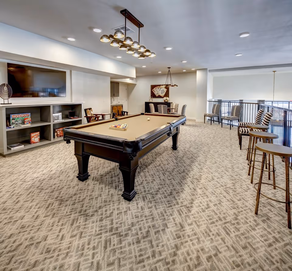 Game room interior with a pool table in the center, a wall-mounted TV and shelving with board games, and multiple seating areas including bar stools and chairs.