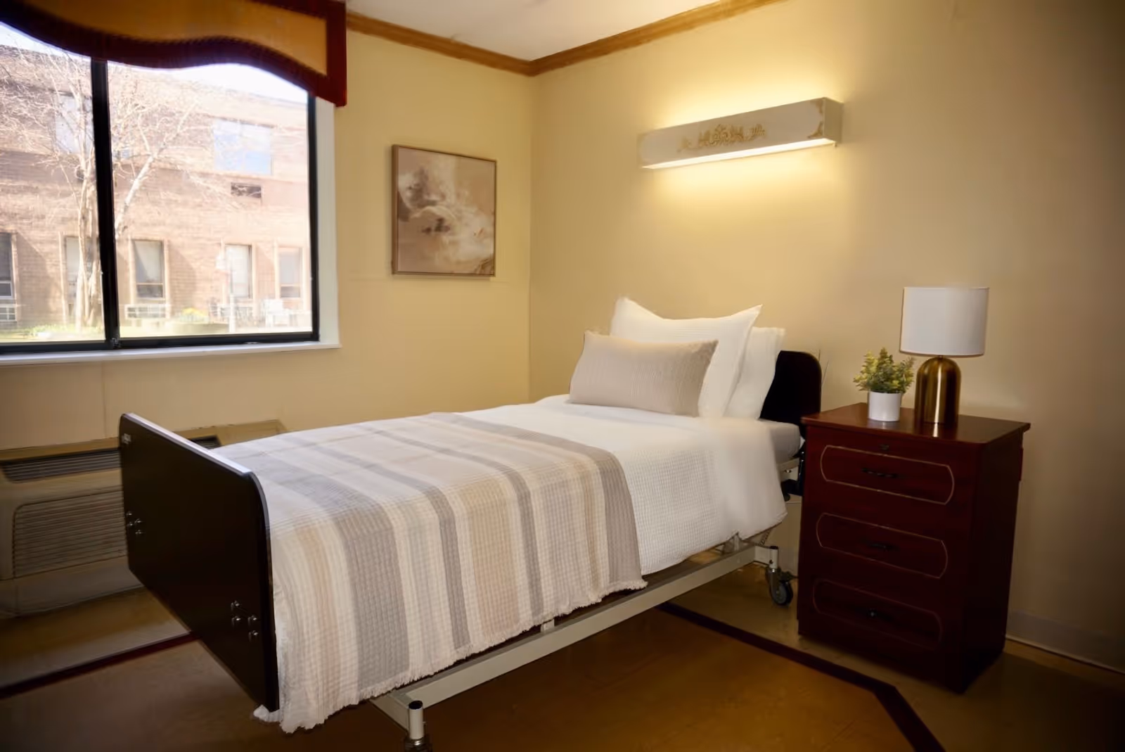 A single hospital-style bed with white and beige bedding in a room with beige walls. Next to the bed is a wooden nightstand with three drawers, a small potted plant, and a lamp with a white shade and gold base. A large window with a dark valance shows an outside view of a brick building and leafless trees. A framed abstract artwork hangs on the wall above the bed, and a wall-mounted light fixture is above the head of the bed.