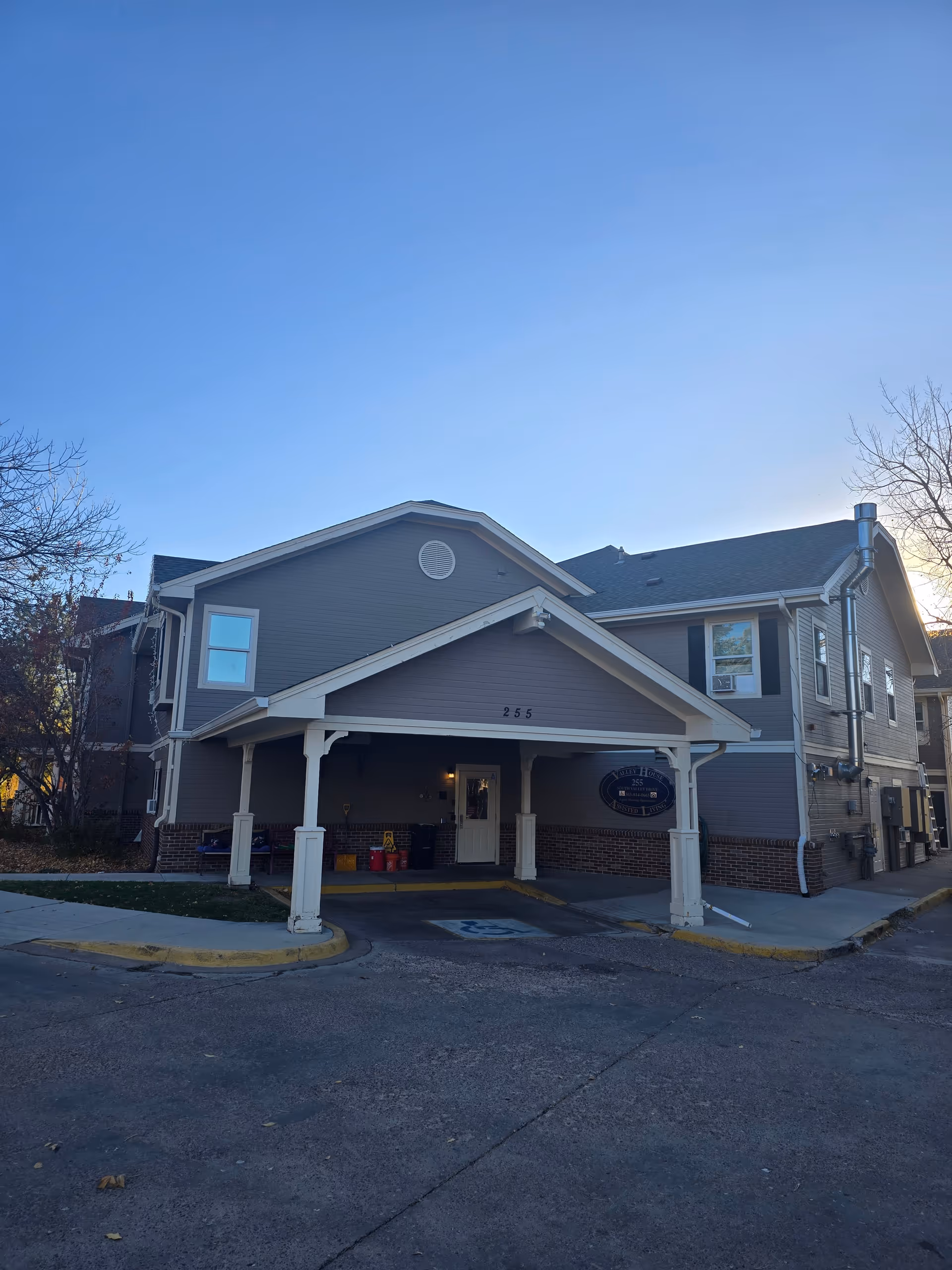 Exterior view of a two-story assisted living facility building with gray siding and white trim. The building has a covered entrance with the number 255 above it and a sign that reads Valley House Assisted Living. There are some trees with autumn leaves around the building and a clear blue sky above.