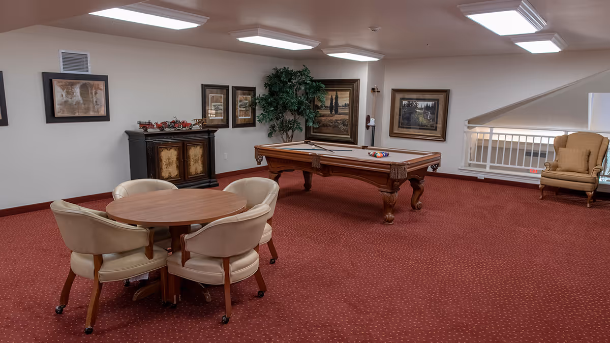 A recreational room with a pool table set up for a game, surrounded by framed artwork on the walls. There is a round wooden table with four beige cushioned chairs on casters, a wooden cabinet with decorative items on top, a large potted plant, and a beige armchair near a white railing overlooking a lower level. The room has red carpet flooring and fluorescent ceiling lights.