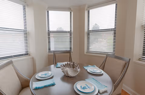 A round dining table set for four with beige cushioned chairs around it, each place setting includes a plate with a blue lobster design and a turquoise napkin. The table has a decorative silver shell centerpiece. The room has three large windows with white blinds, allowing natural light to enter.
