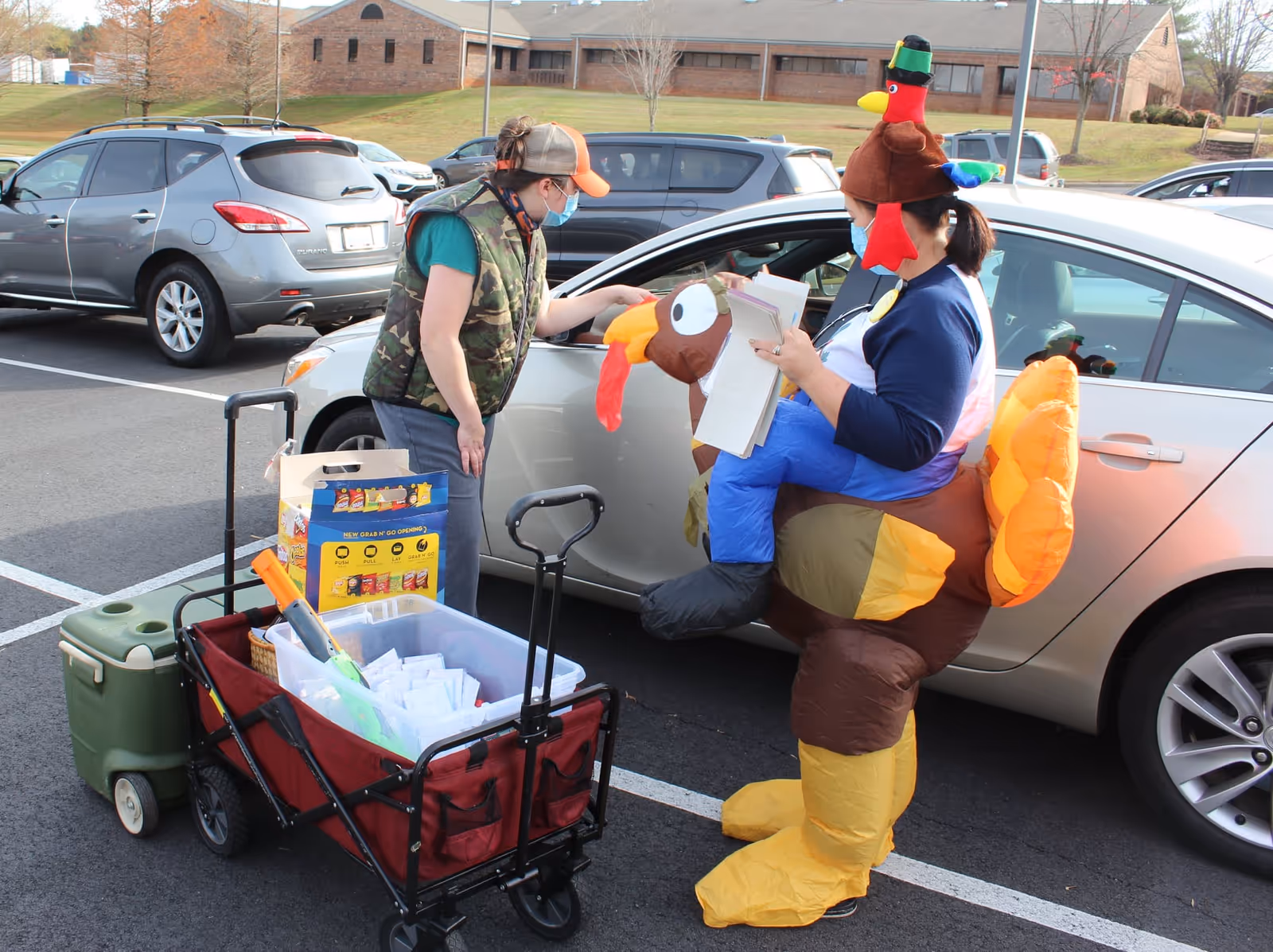 A person wearing a colorful inflatable turkey costume and a face mask is handing papers to another person wearing a camouflage vest, orange cap, and face mask. They are standing next to a silver car in a parking lot with other parked cars and a brick building in the background. There is a red wagon filled with supplies and a green cooler nearby.
