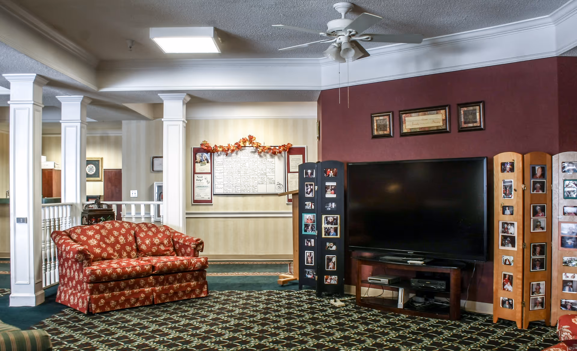Communal living room with a red floral loveseat, patterned carpet, a large flat-screen TV flanked by photo display panels, and a ceiling fan.
