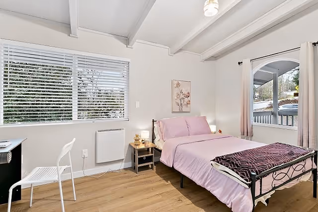 A bright, simply furnished bedroom with a pink-made bed, nightstand, desk, wood floor and large windows beneath a white beamed ceiling.