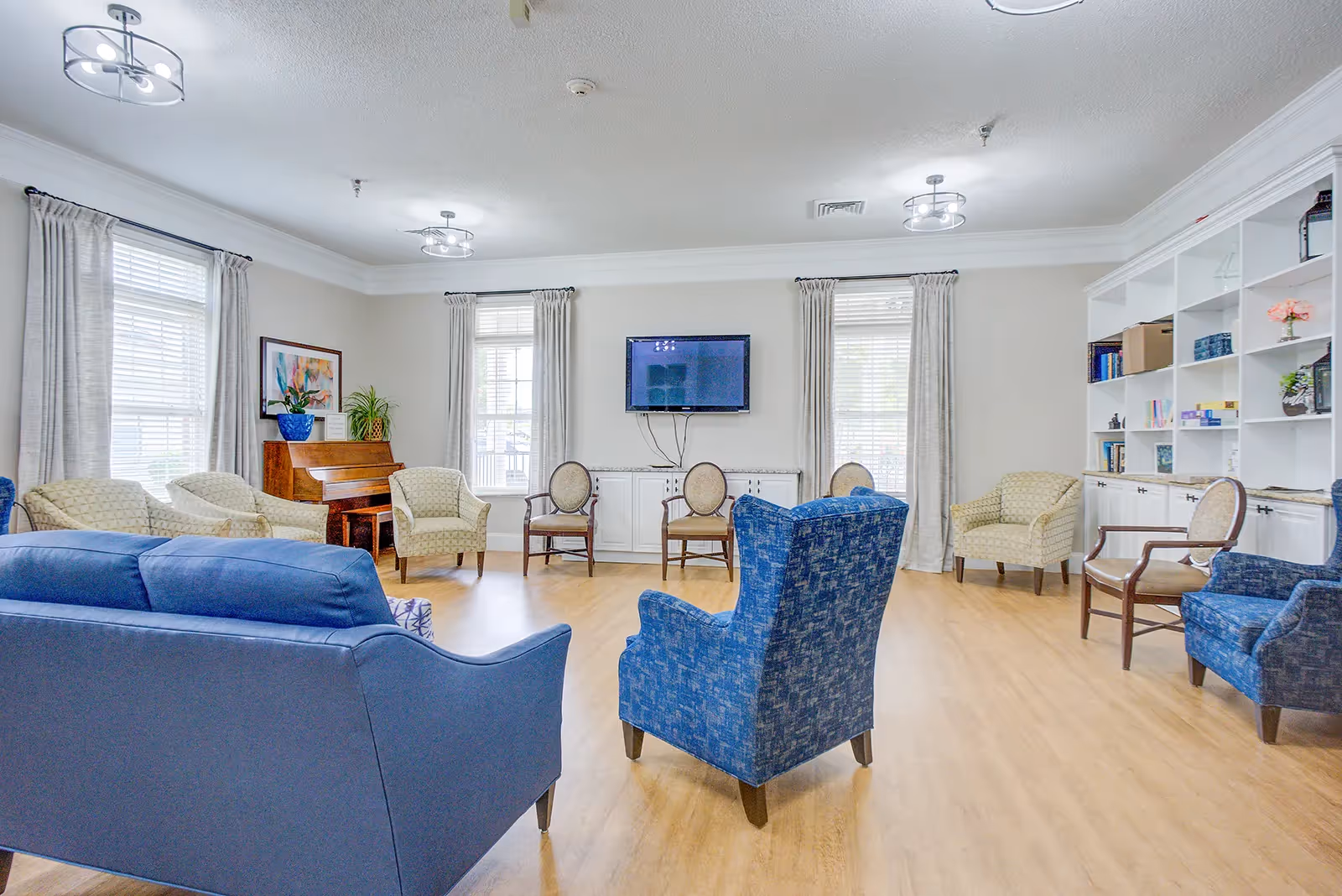 Bright senior living common room with sofas and armchairs arranged around a wall-mounted TV and a piano.