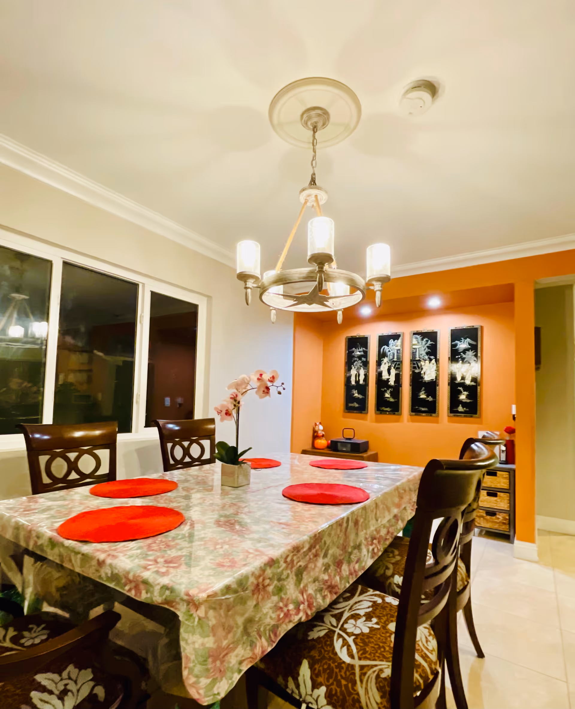A dining room with a rectangular table covered with a floral tablecloth and a clear plastic cover. The table has six wooden chairs with patterned cushions and red round placemats. A small potted orchid is placed in the center of the table. Above the table hangs a chandelier with four lights. The walls are painted white and orange, with four black decorative panels on the orange wall. There is a window on the left side and a small cabinet with drawers on the right side.