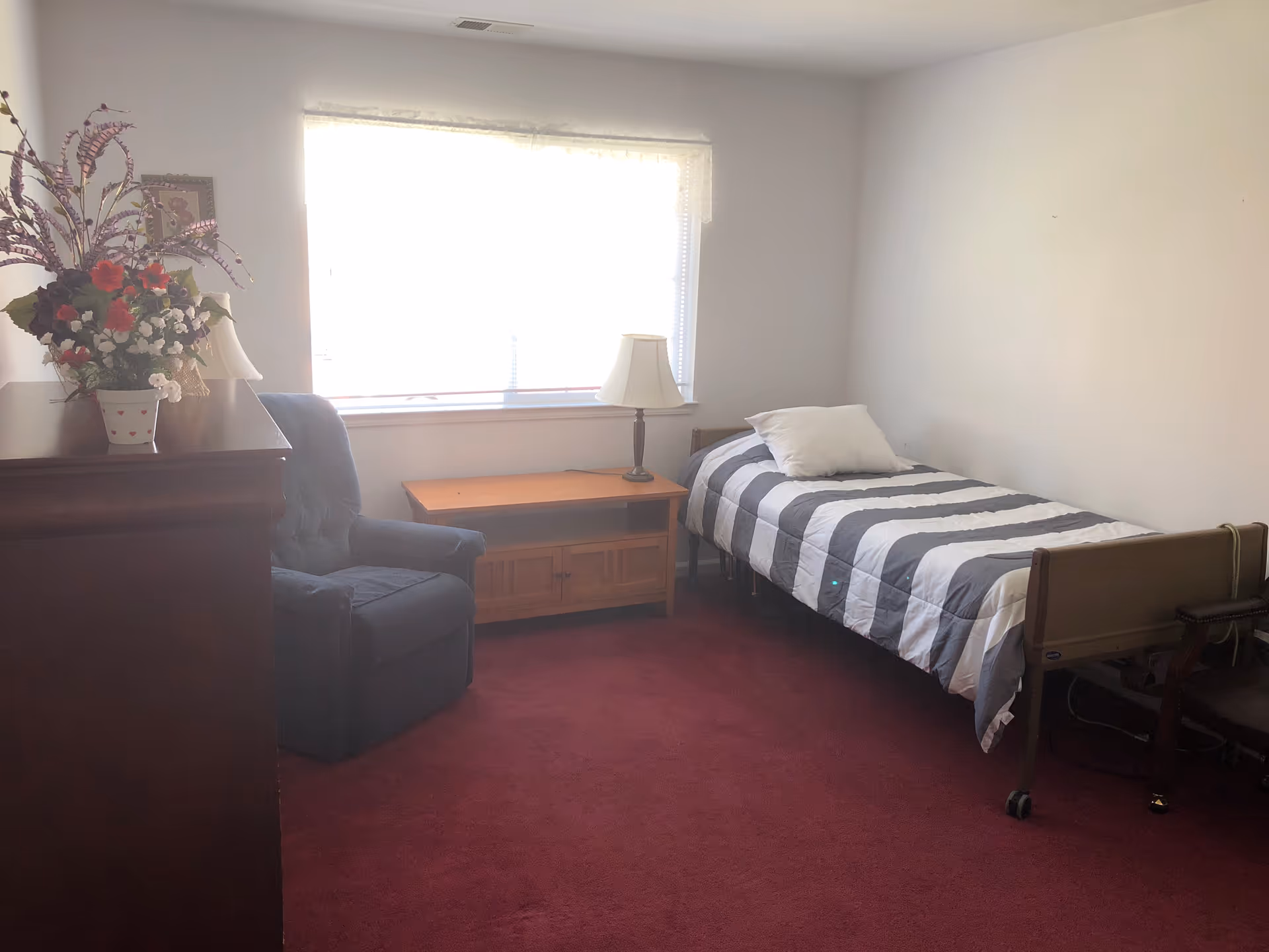 A small bedroom with a single bed covered in a gray and white striped comforter and a white pillow. Next to the bed is a wooden nightstand with a lamp on it. There is a blue upholstered armchair beside a wooden dresser with a floral arrangement on top. The room has a large window with blinds and a red carpeted floor.