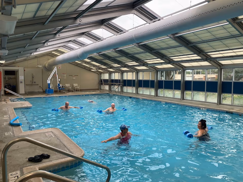 Indoor swimming pool with four people exercising in the water using blue foam dumbbells. The pool area has a high ceiling with large windows along one side, allowing natural light to enter. There are metal handrails and steps leading into the pool, and some chairs and pool equipment are visible in the background.