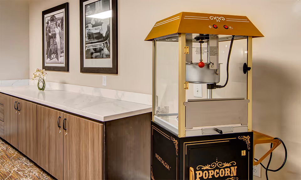 A popcorn machine labeled 'Nostalgia' and 'Popcorn' sits next to a long countertop with wooden cabinets below and a white marble-like surface. Two black and white framed photographs hang on the beige wall above the countertop, and a small vase with flowers is placed on the counter.