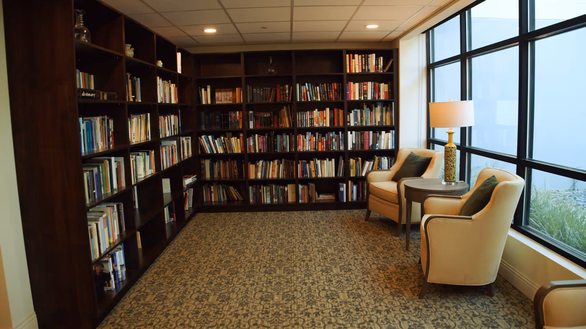 Cozy reading room with wall-to-wall bookshelves, two armchairs and a side table with a lamp beside large windows.