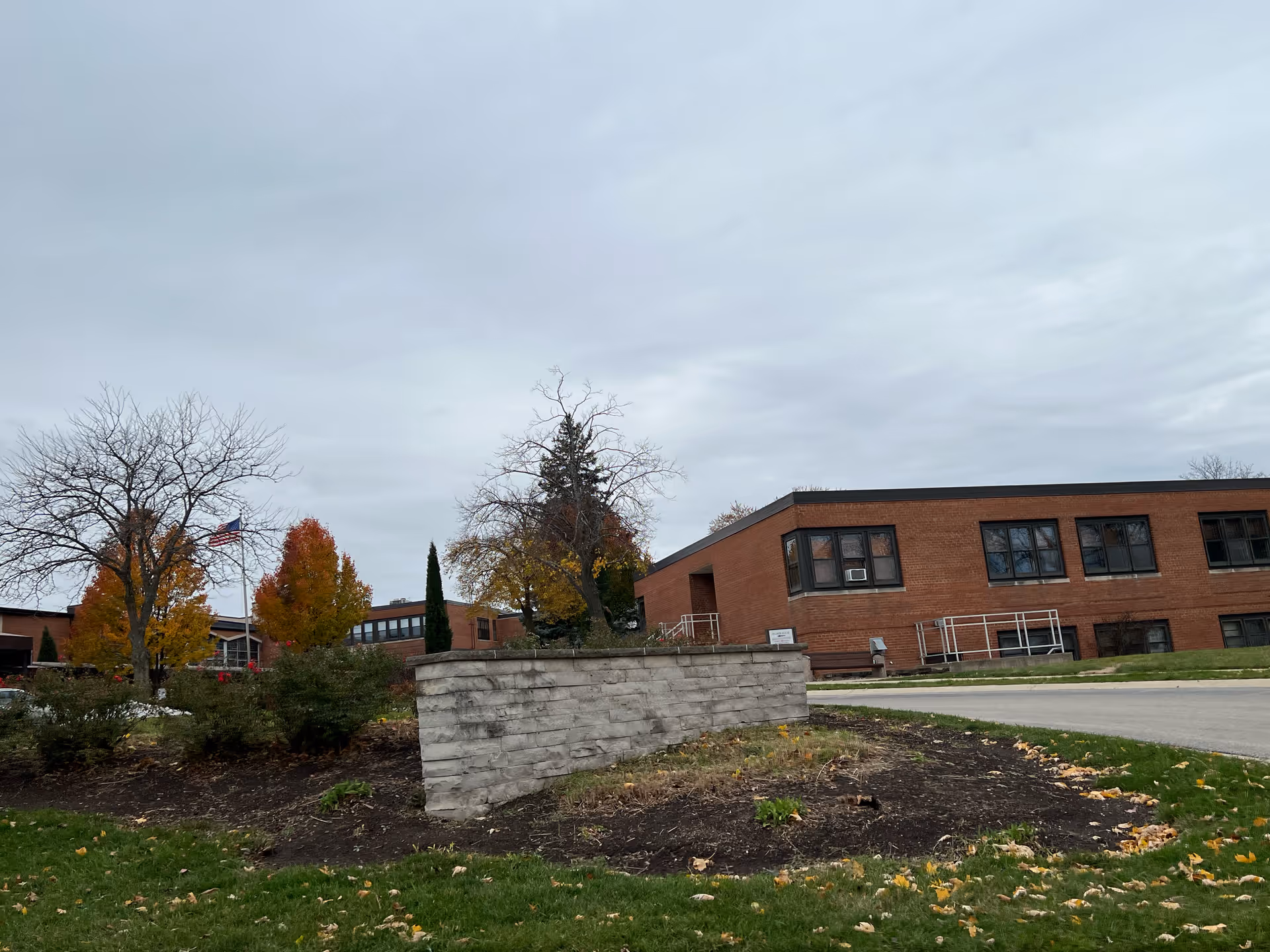 Brick senior living facility building with a low stone planter, autumn trees, and an overcast sky.