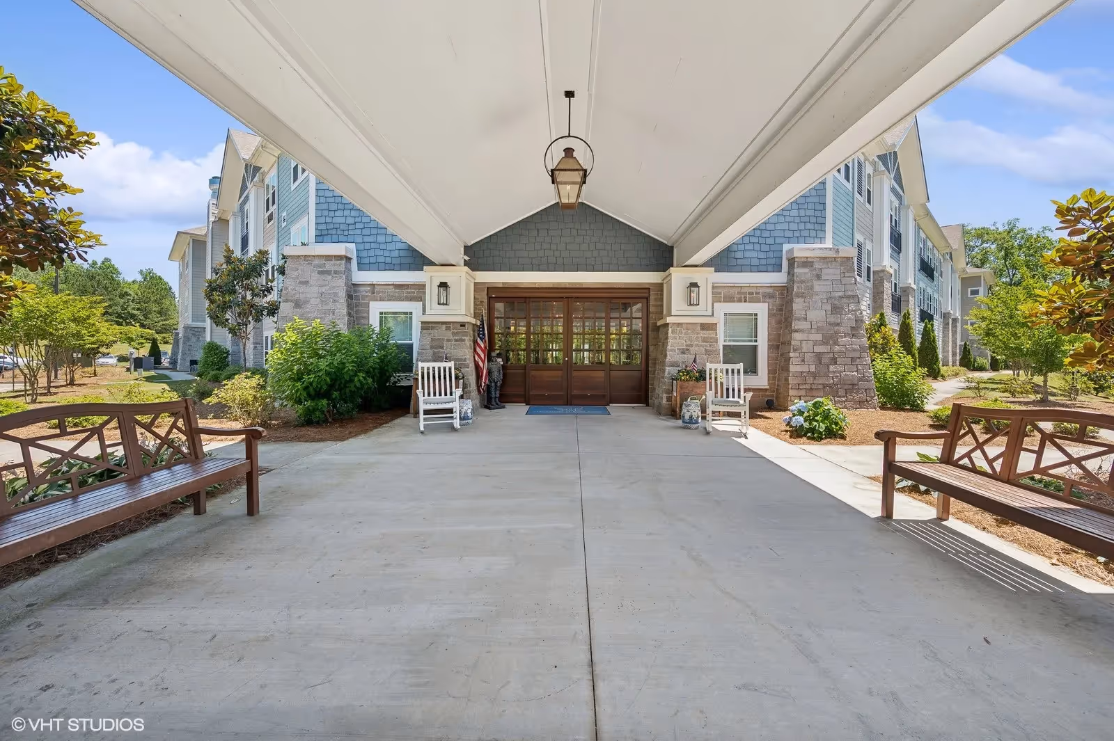 Covered entrance/porte-cochere of a senior living building with benches, rocking chairs, landscaping, and glass double doors.