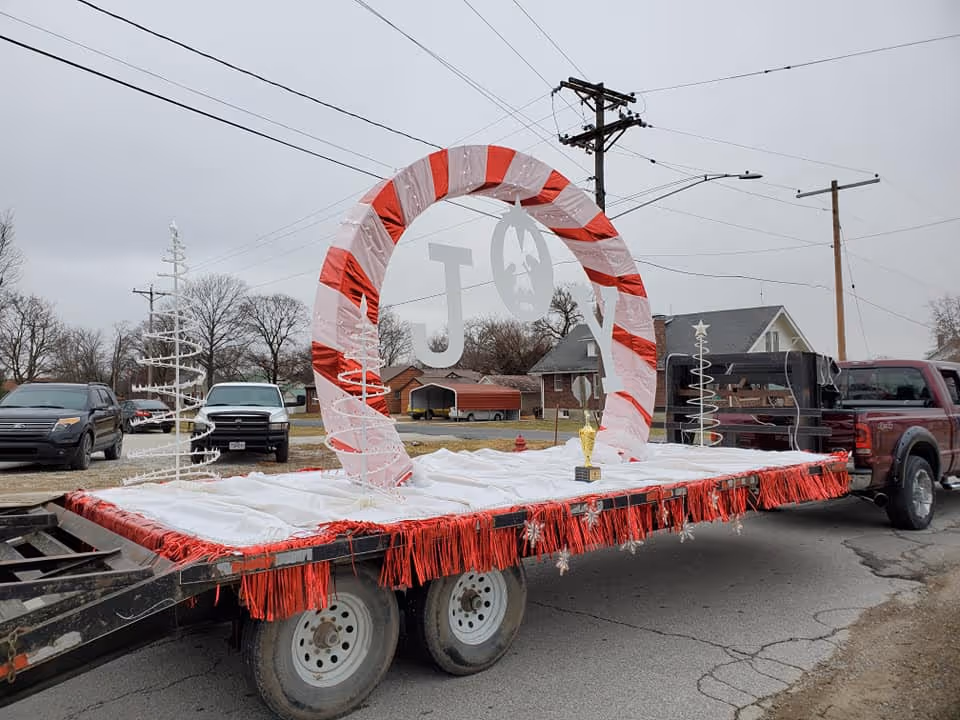A pickup truck towing a decorated parade float with a red-and-white candy-cane arch spelling 'JOY' parked on a residential street.